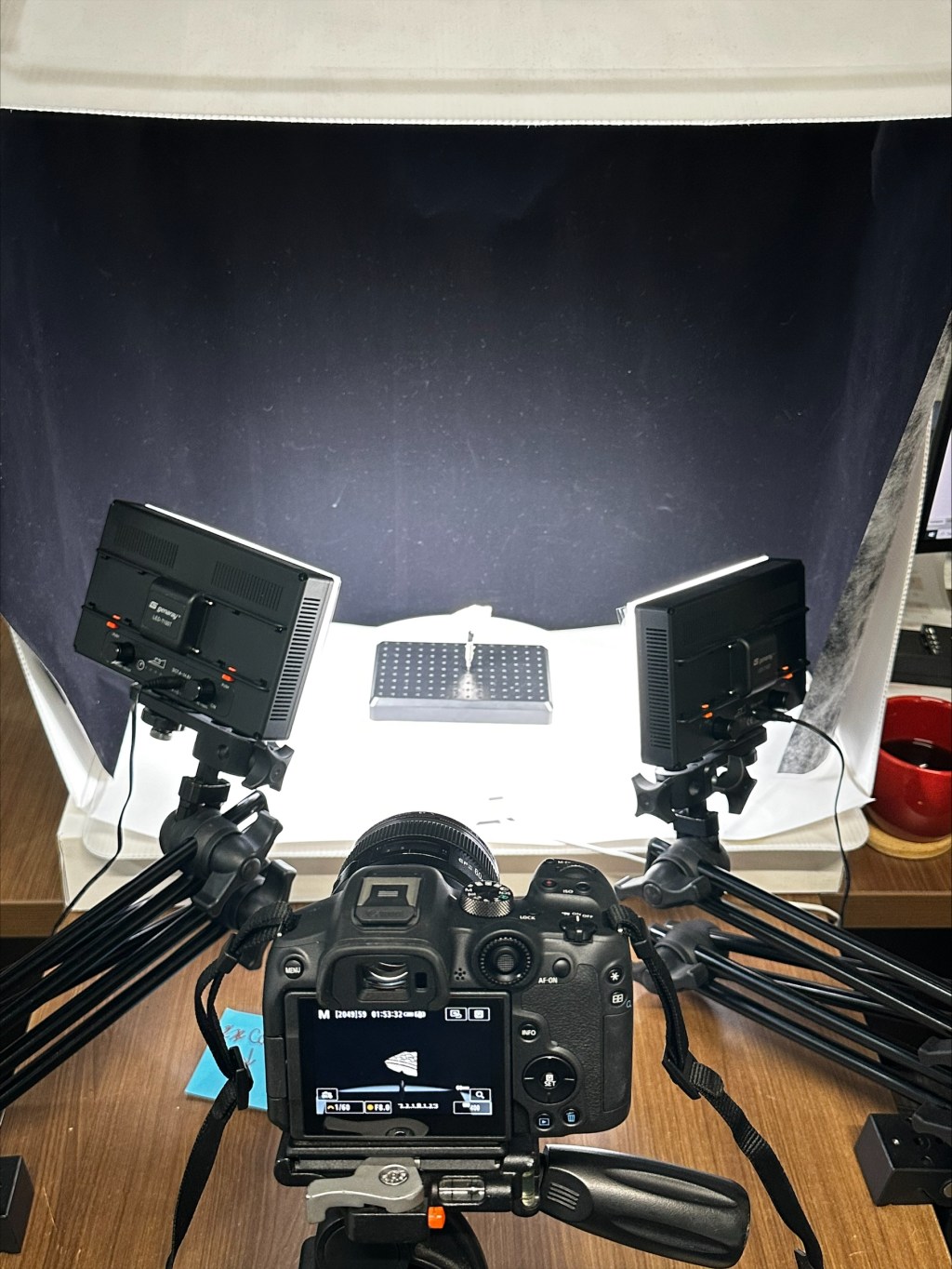 A digital camera stands on a tripod atop a desk, pointed at an artifact on a turntable. Lights on each side illuminate the object.